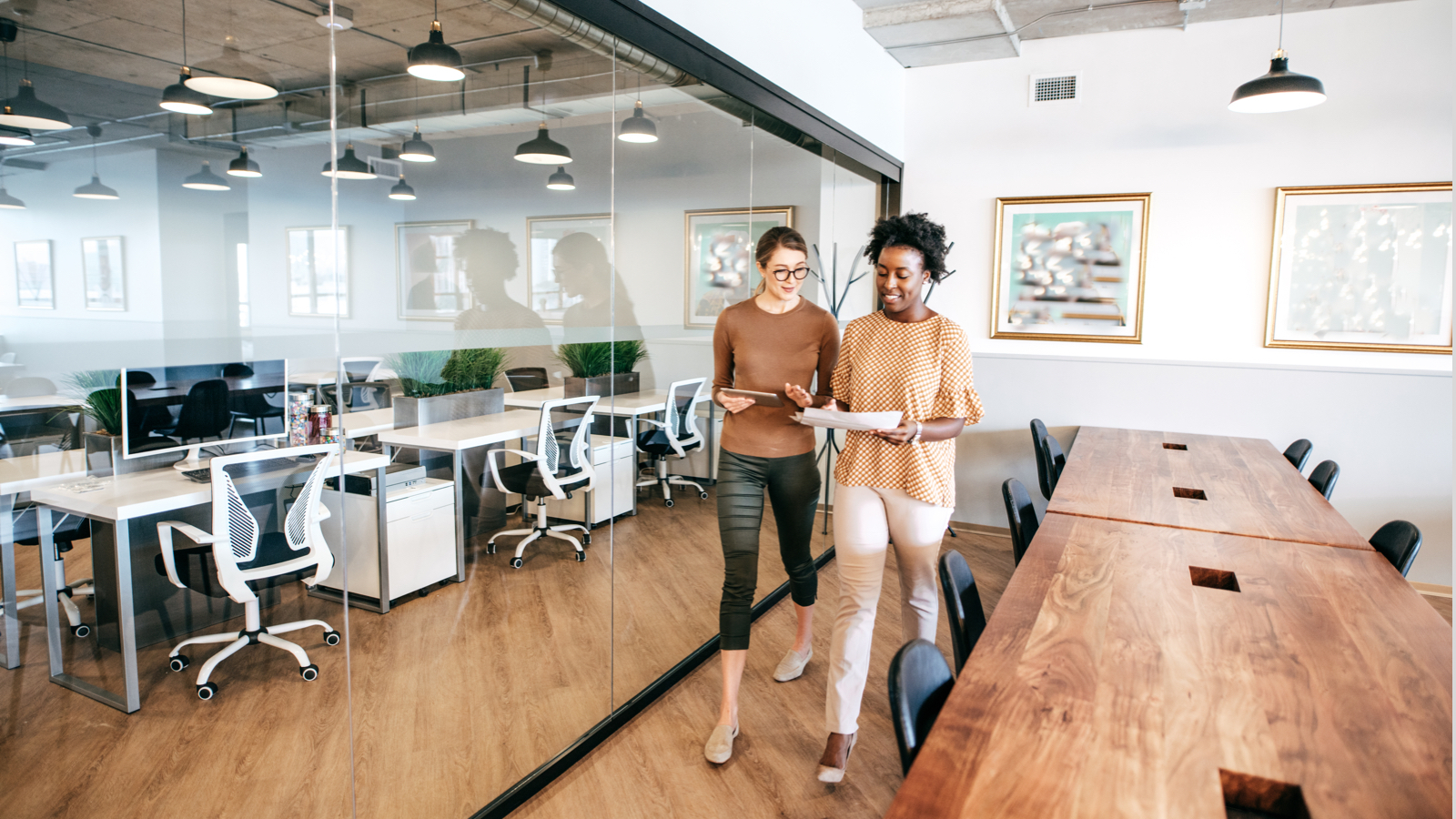 Two professional women leaving conference room in a brightly lit, open space office.