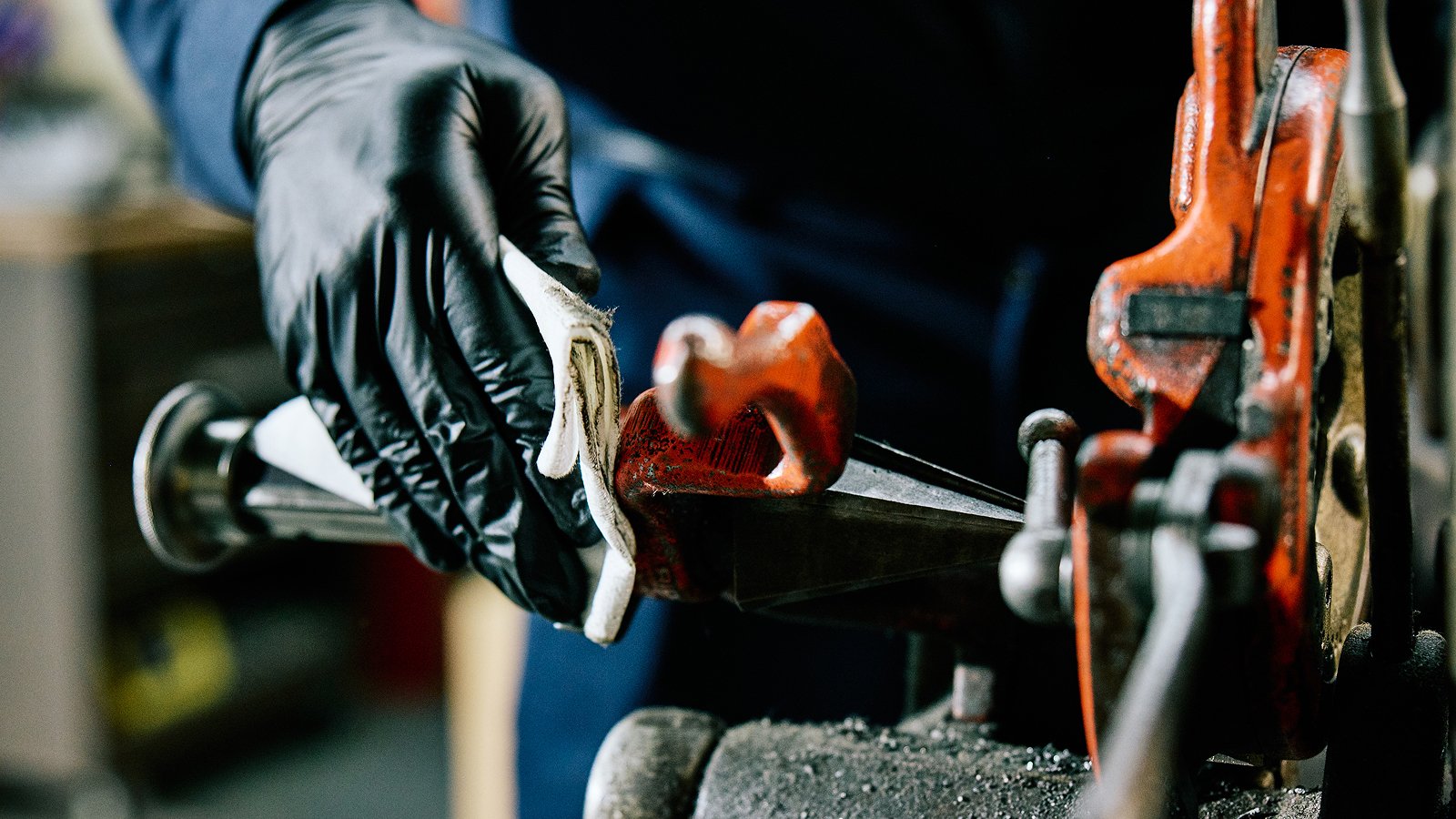 - Person wearing black gloves using a WypAll® X80 heavy-duty cleaning cloth to clean a metal surface, with the cloth absorbing liquid.