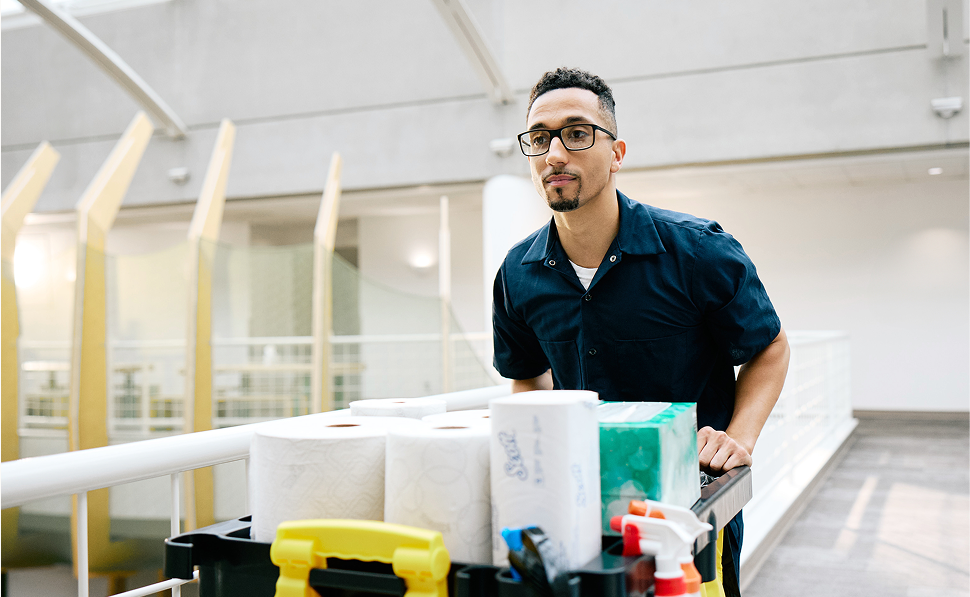 A professional cleaner pushing a cart filled with hygiene products, including paper towels, in a modern indoor setting. 